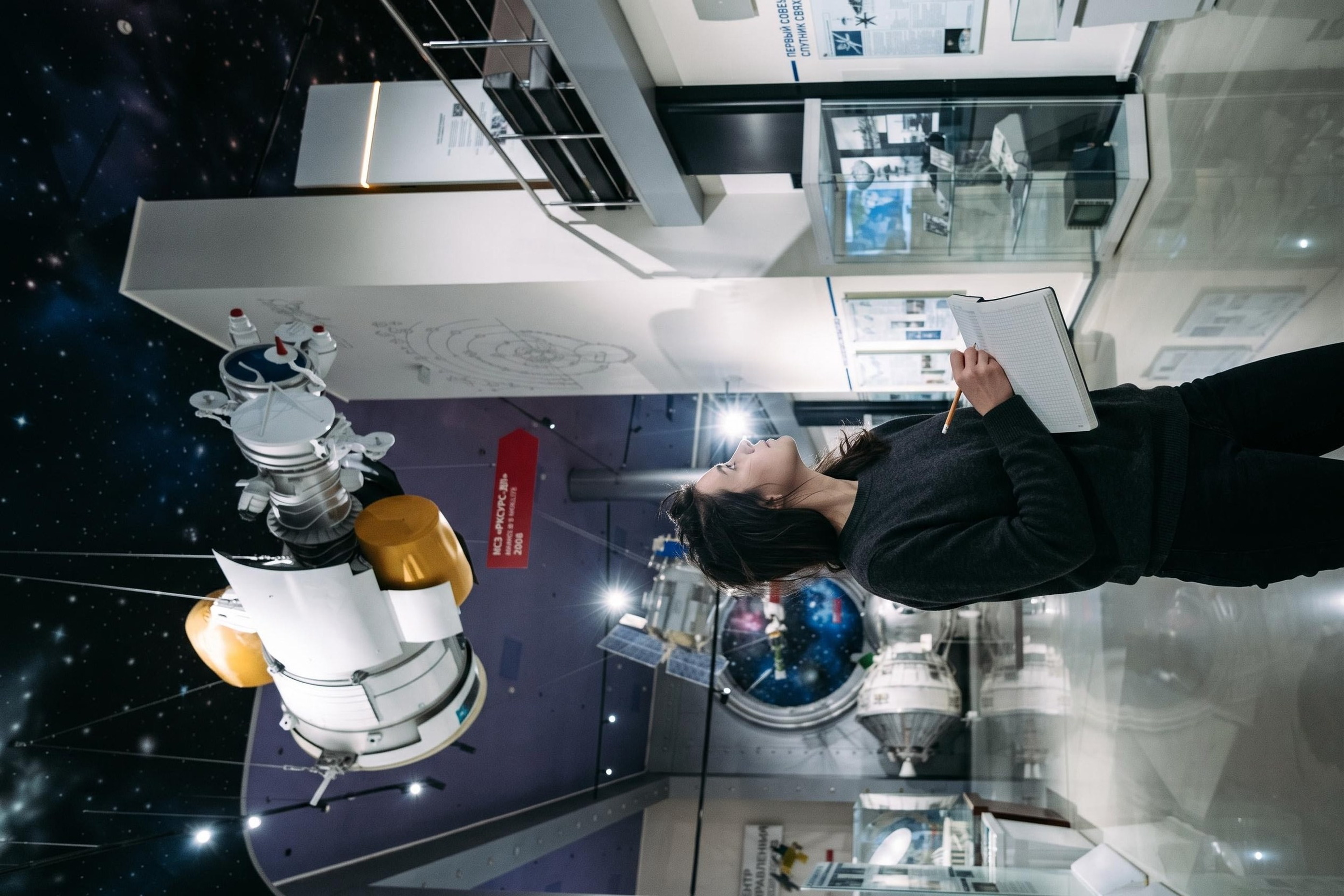 Me below a suspended satellite in the Museum of Cosmonautics, notebook open on my forearm.