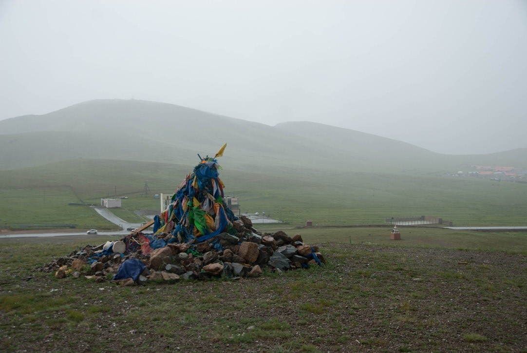 Stone ovoo with weathered blue prayer scarves on a rainy green pass.