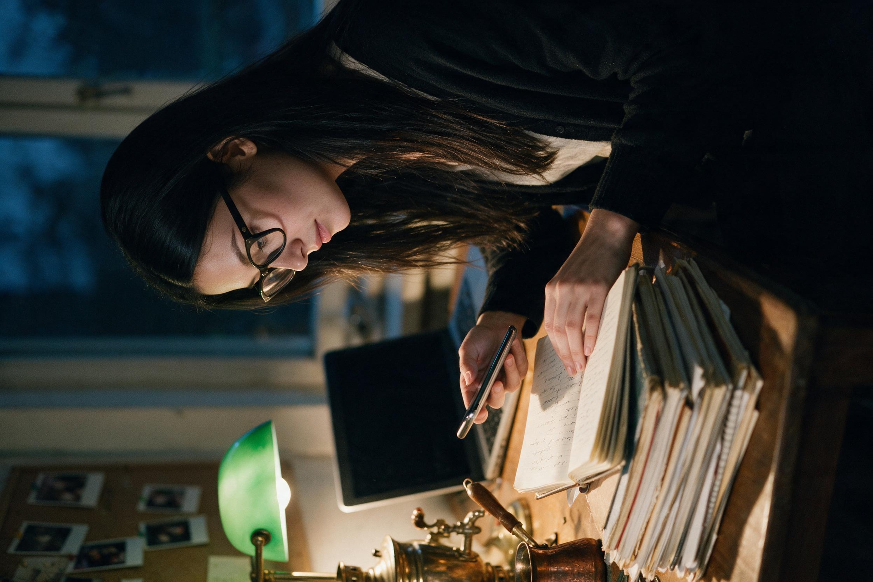Me at the desk at night, photographing a page of an old notebook with my phone, the rest of the stack waiting under it.
