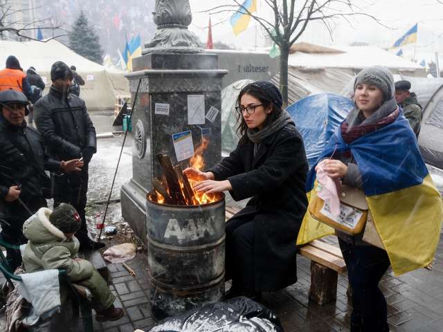 Me at a fire barrel with others at the memorial camp. Someone in a Ukrainian flag nearby.
