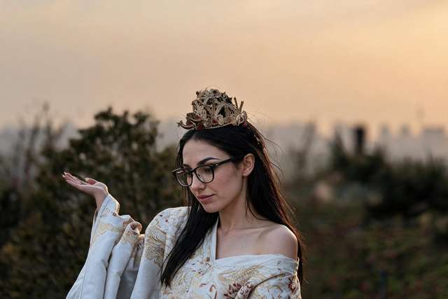 Me in a white and gold phoenix robe and wedding crown at sunset, one shoulder bare, a leaf on my hand.