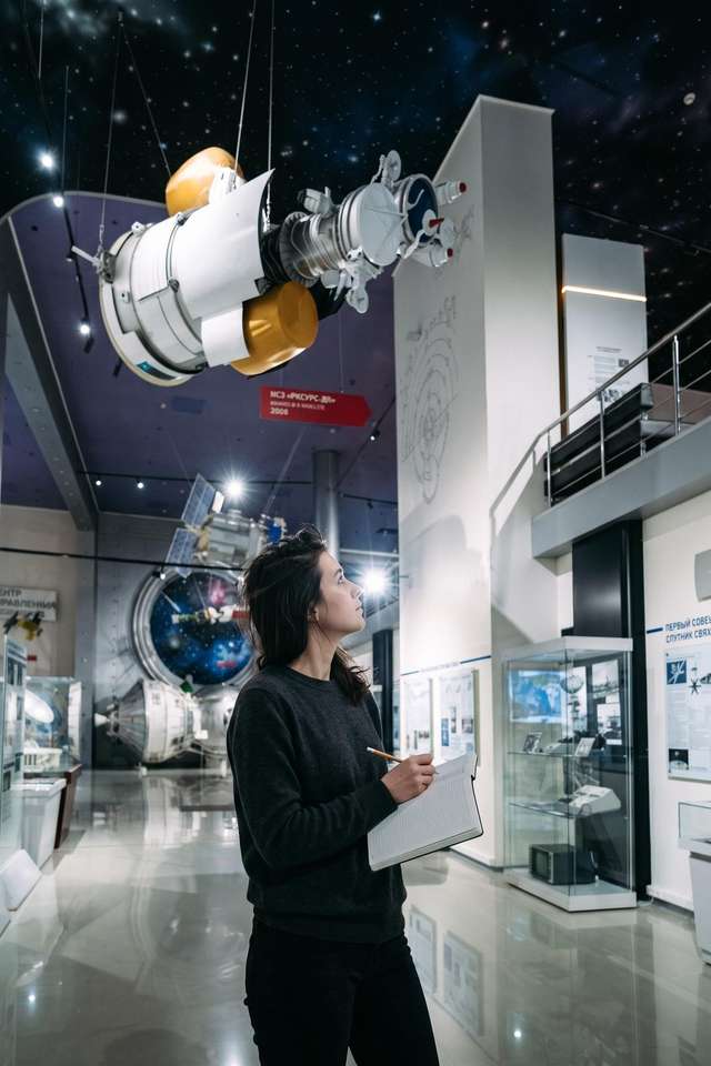 Me below a suspended satellite in the Museum of Cosmonautics, notebook open on my forearm.