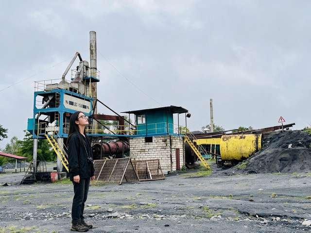 Me with a camera before an abandoned blue asphalt plant and its rusty silo.