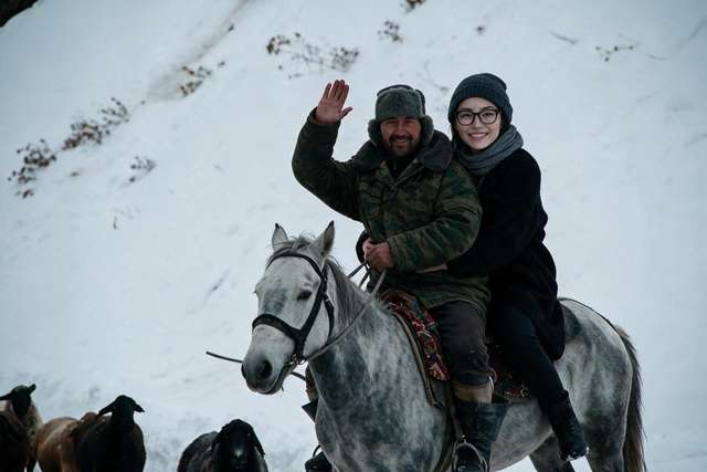 Me grinning behind a herder in a fur hat on his white horse in snow. Sheep ahead.