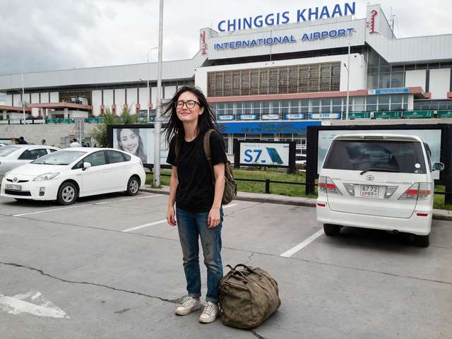 Me with my duffel in front of the airport terminal, tired smile.