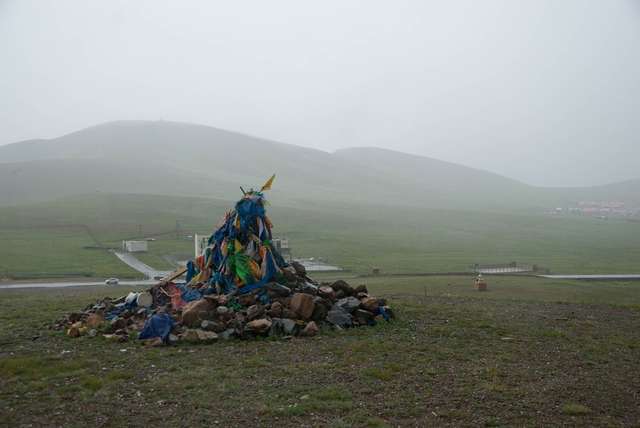 Stone ovoo with weathered blue prayer scarves on a rainy green pass.