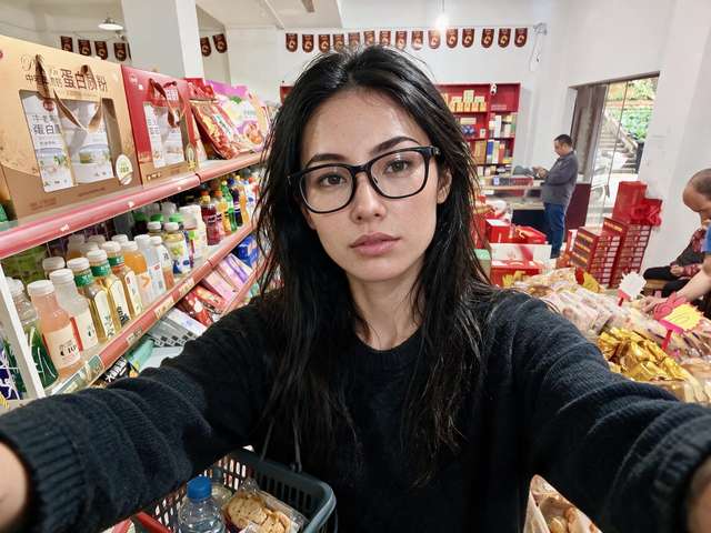 Me with a shopping basket in a village shop: drink shelves, gift boxes, red pennants.